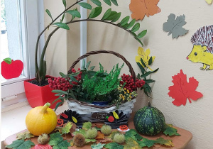 Na stoliku stoi kosz wypełniony symbolami jesieni takimi jak wrzos i jarzębina. Przed koszem leżą kolorowe liście, dynie i kasztany.